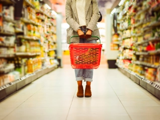 A shopper holding a red plastic shopping basket stands in the brightly lit aisle of a supermarket between two long, stocked shelves.