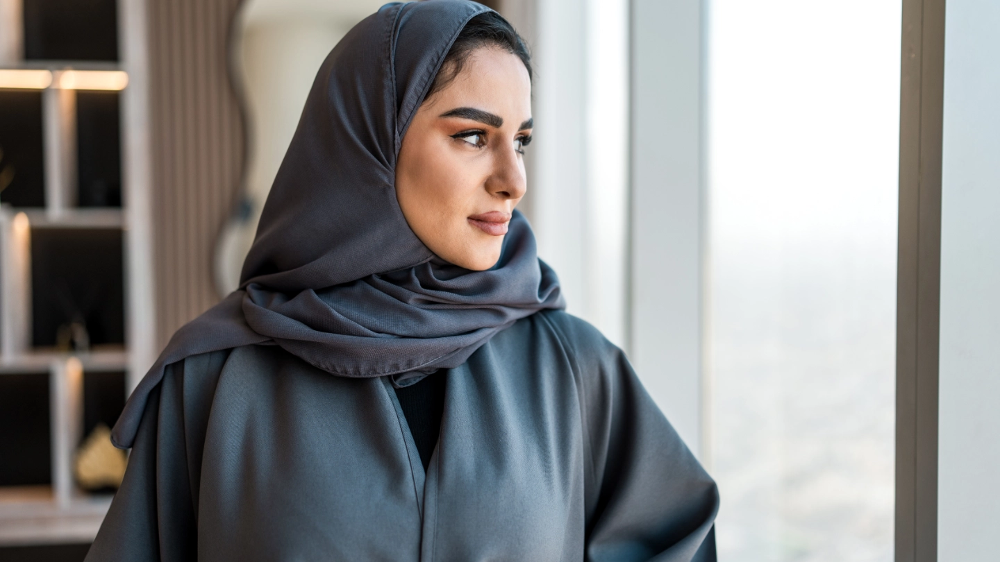 An Arabic woman wearing a gray abaya and matching hijab looks thoughtfully out of a large window with a city view.